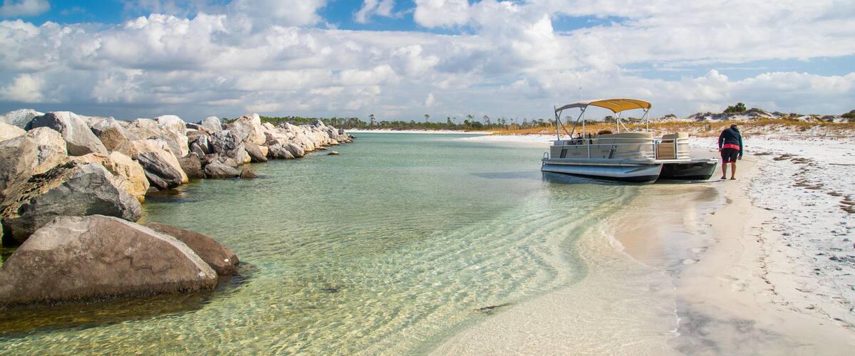 A clear turquoise sea with a boat docked on a sandy beach, surrounded by large rocks and palm trees under a partly cloudy sky.