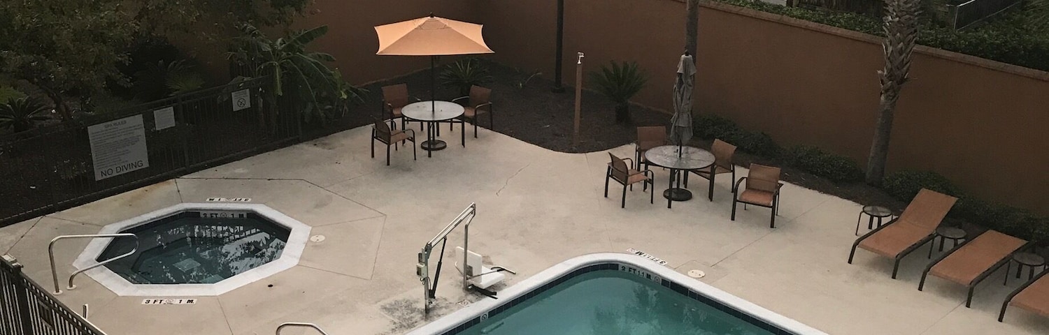 A swimming pool with lounge chairs and a hot tub area, surrounded by a fence and palm trees.