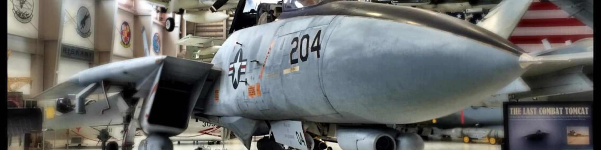 A military aircraft with the number 204 displayed on its nose, parked indoors with other aircraft and a display board in the background.