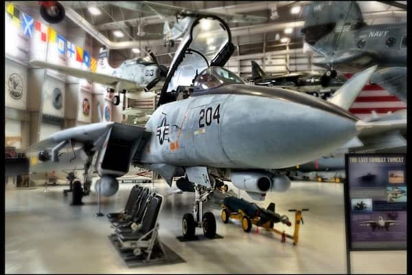 Ein Militärflugzeug mit der Aufschrift 204 auf der Nase, geparkt in einem Innenbereich, mit weiteren Flugzeugen und einer Werbetafel im Hintergrund.