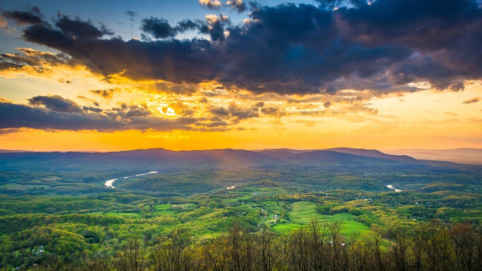 A valley with a river, surrounded by green hills and a dramatic sky with clouds.