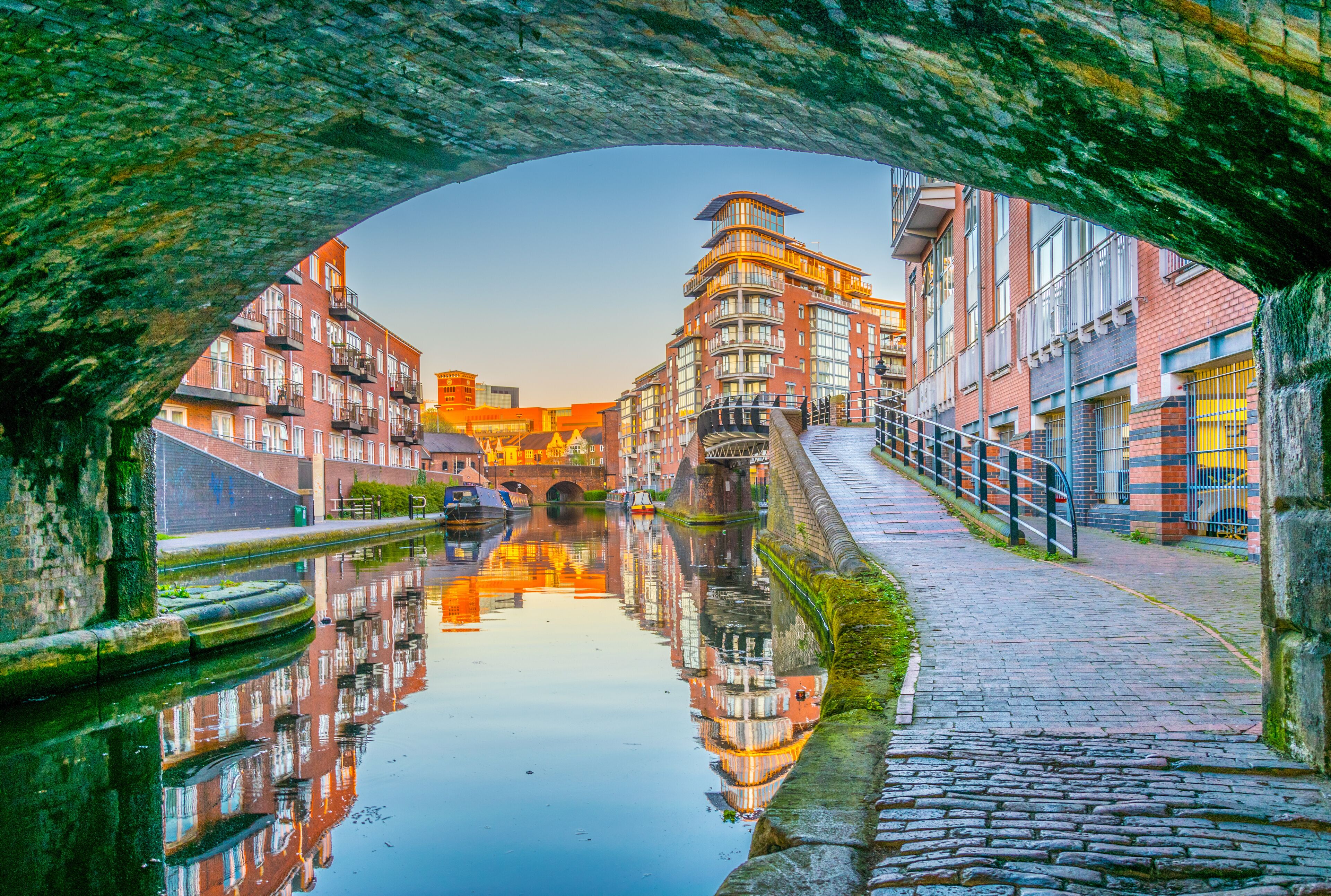 A canal with buildings reflected in the water, a stone bridge, and a cobblestone path.