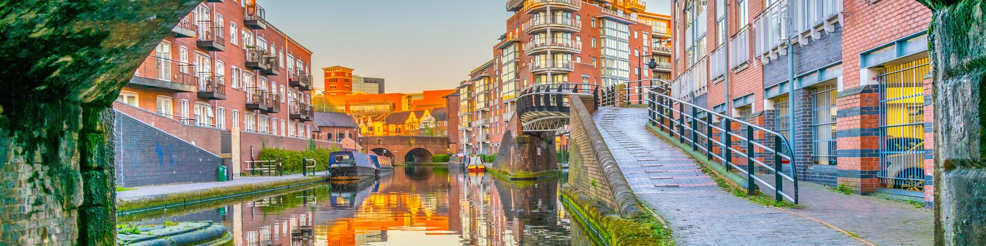 A canal with buildings reflected in the water, a stone bridge, and a cobblestone path.