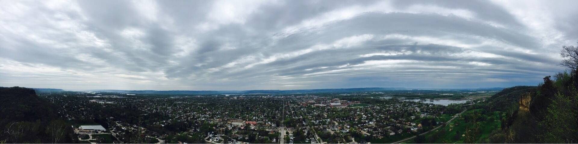 A panoramic view of a city with a river, surrounded by hills and a cloudy sky.