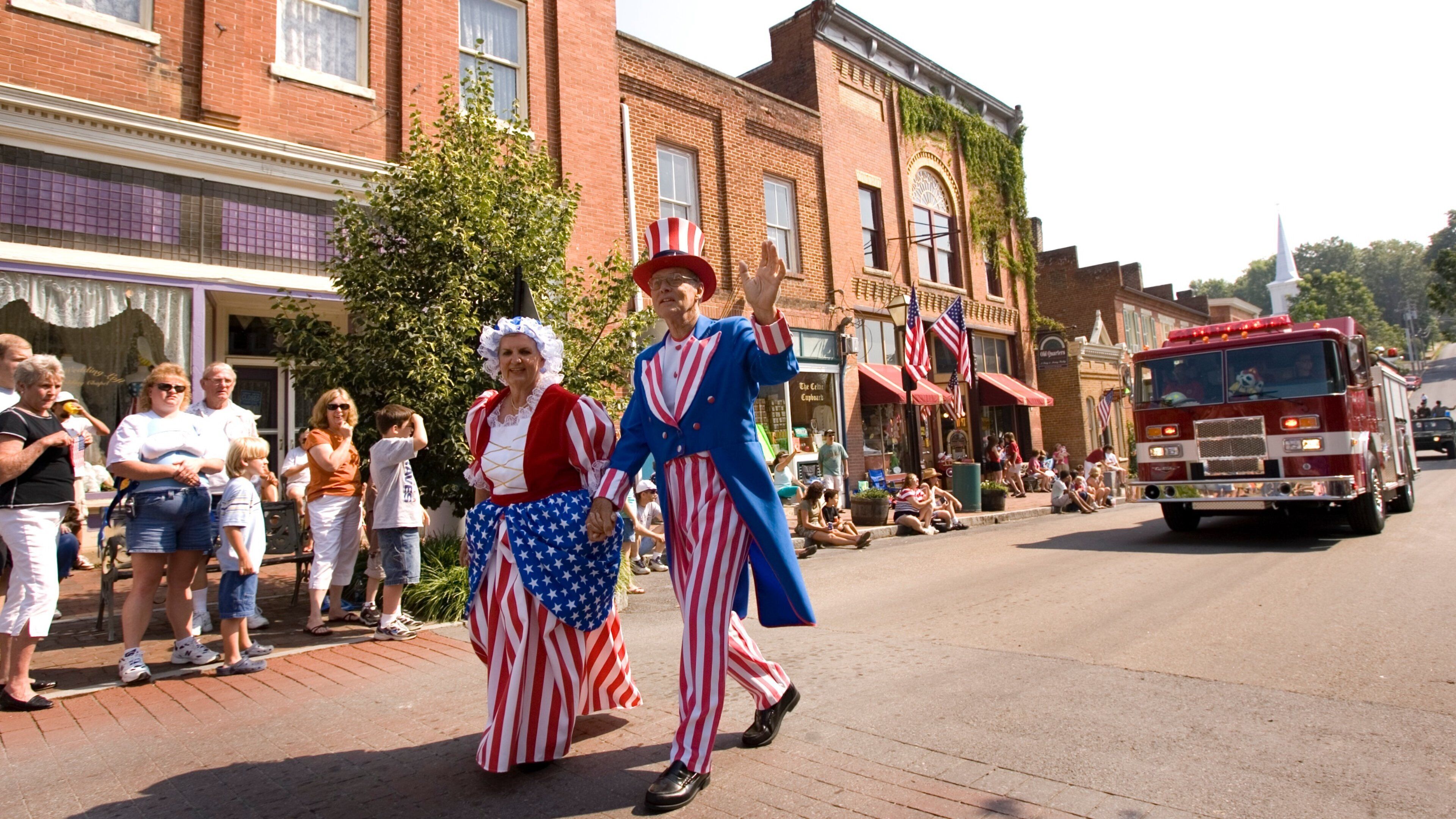 A parade with people in patriotic costumes, a fire truck, and brick buildings.