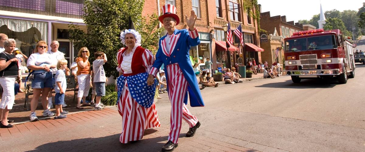 A parade with people in patriotic costumes, a fire truck, and brick buildings.