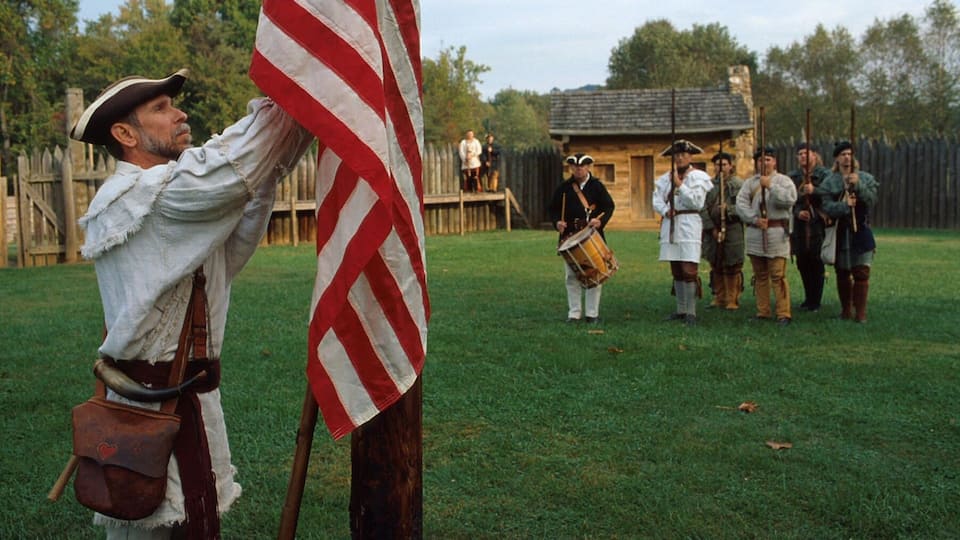 A historical reenactment scene with a person in colonial attire holding an American flag.