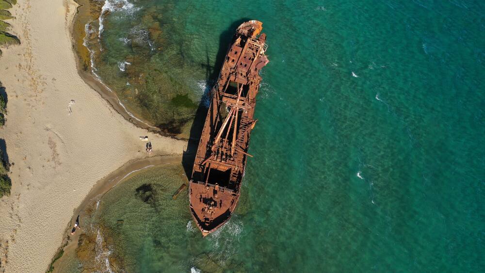 An aerial view of a beach with a rusted shipwreck partially submerged in clear turquoise waters.