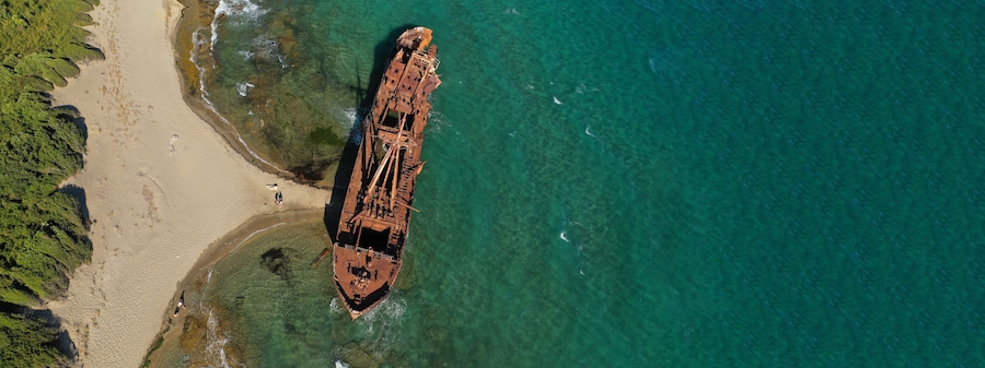Une vue aérienne d’une plage où l’on aperçoit l’épave rouillée d’un navire, partiellement immergée dans des eaux turquoise et cristallines.