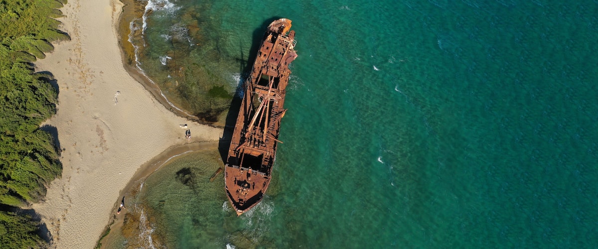 An aerial view of a beach with a rusted shipwreck partially submerged in clear turquoise waters.