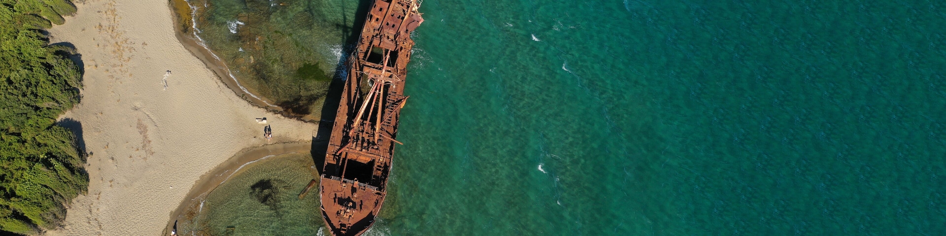An aerial view of a beach with a rusted shipwreck partially submerged in clear turquoise waters.