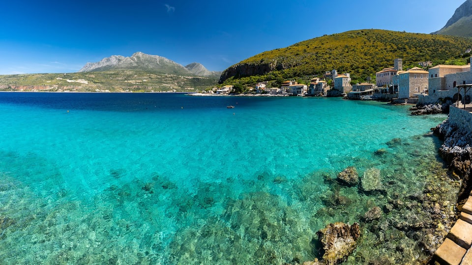 A coastal village with clear turquoise waters, stone buildings, and a mountain in the background.