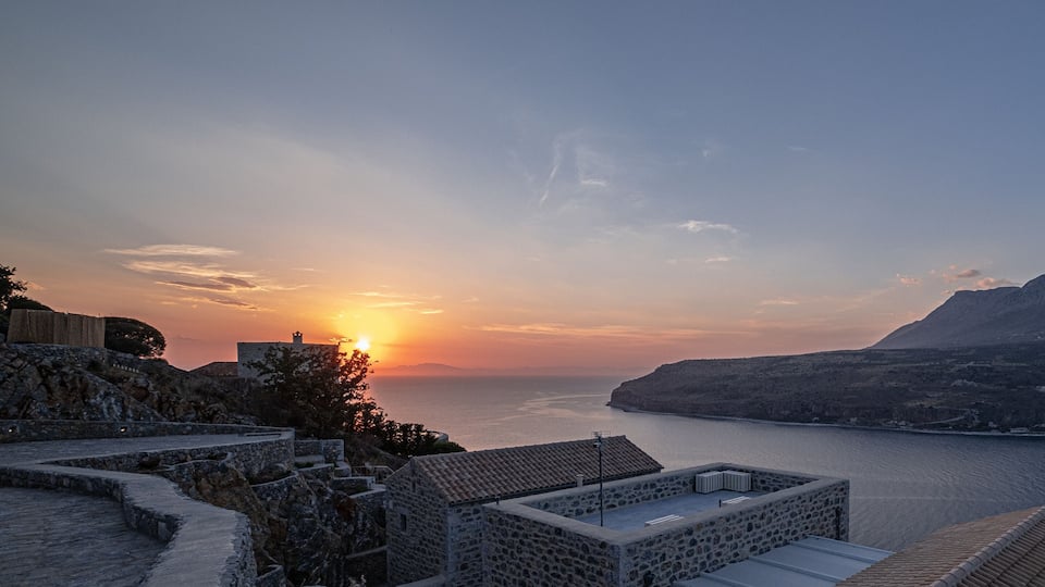 A coastal sunset with stone buildings and a mountainous backdrop.