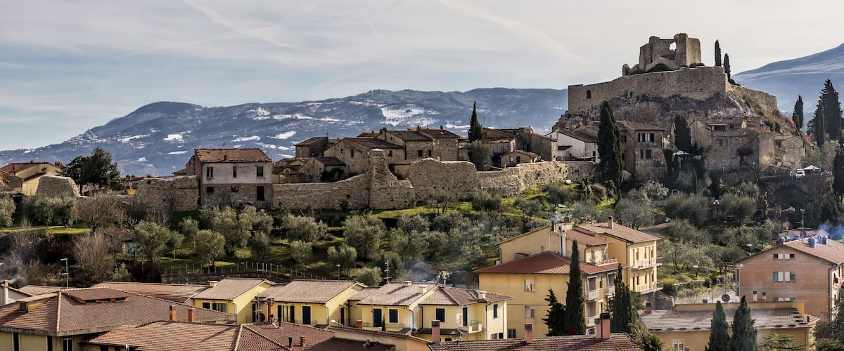 A historic town with stone buildings, a prominent castle on a hill, and snow-capped mountains in the background.