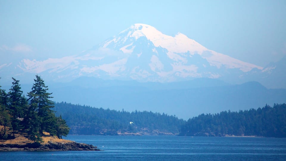 A mountain with snow-capped peaks, a forested shoreline, and a calm body of water.