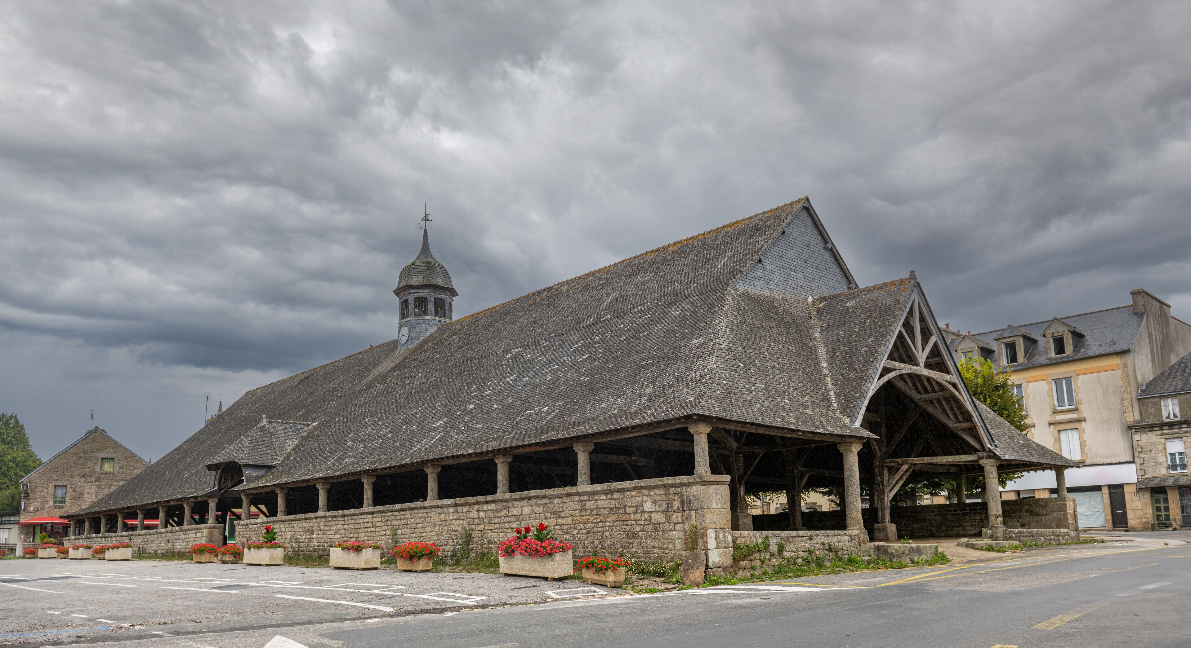 A historic stone building with a steep roof and a bell tower, surrounded by flower beds and a paved street.