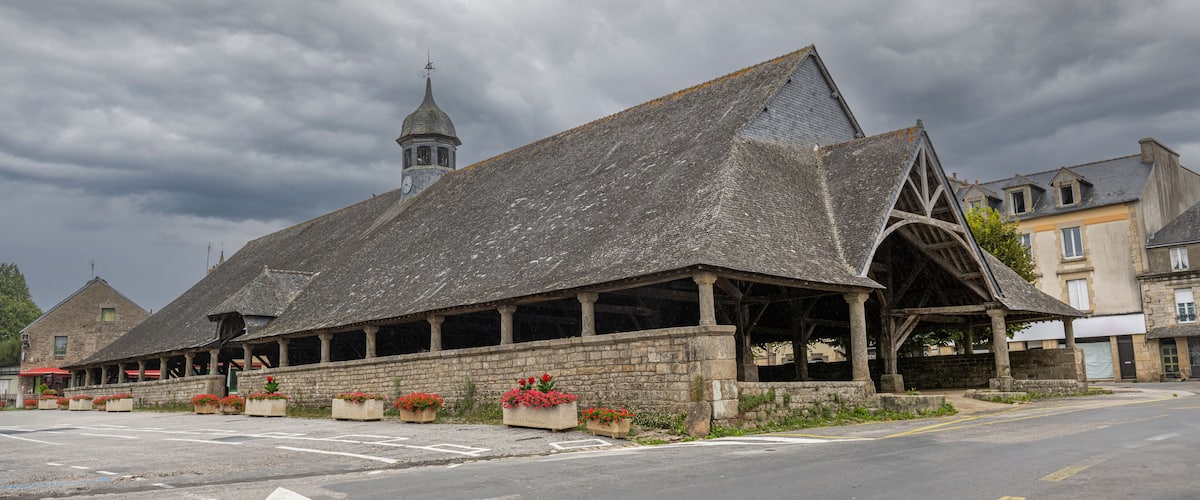 A historic stone building with a steep roof and a bell tower, surrounded by flower beds and a paved street.