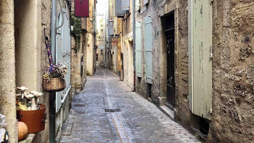 A narrow cobblestone street flanked by old buildings with shutters and a basket of flowers hanging on the wall.