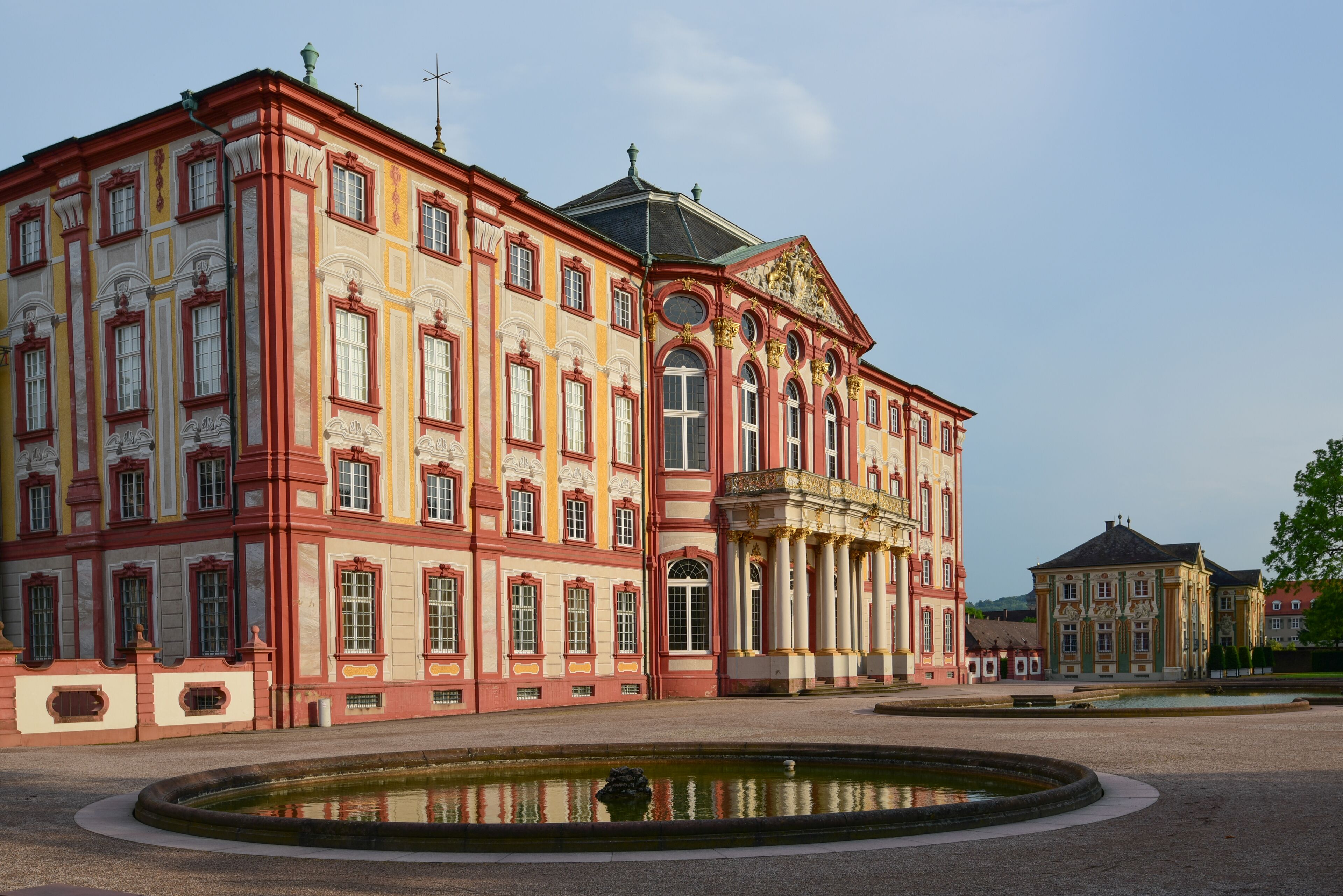 A large, ornate building with red and yellow facades, a central dome, and a surrounding courtyard with a fountain.