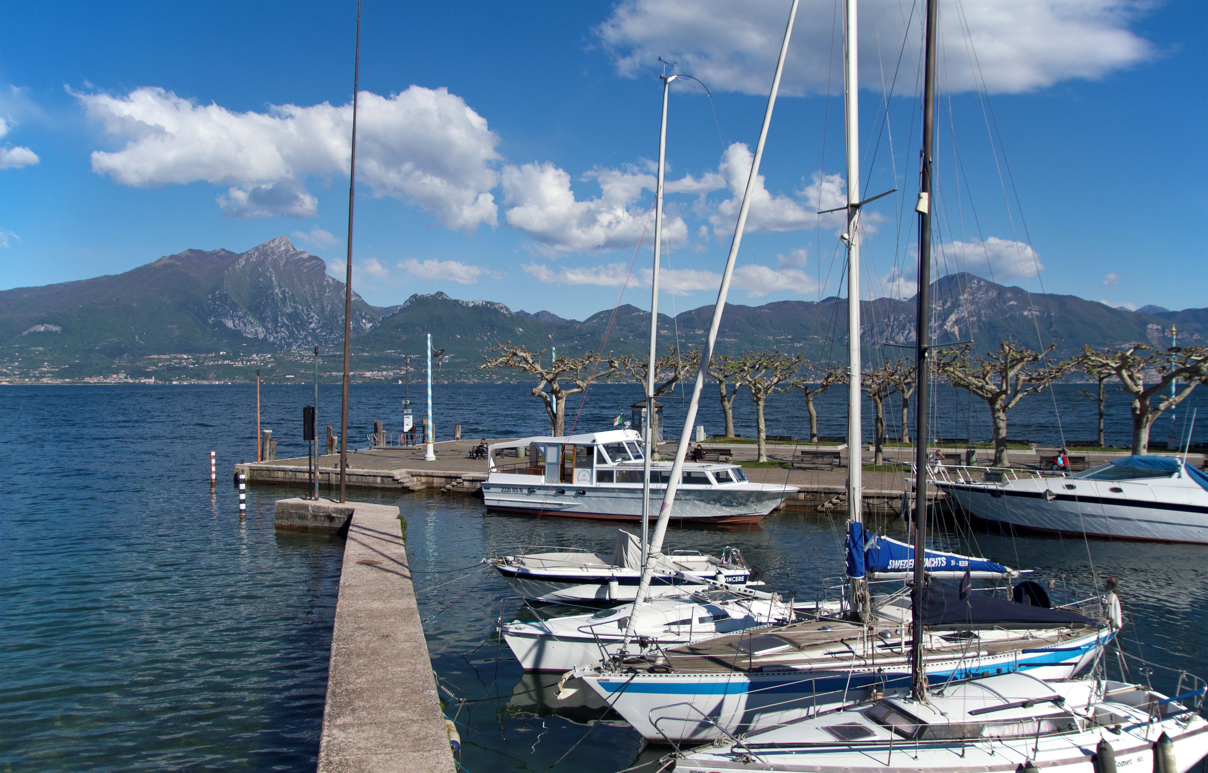 A marina with several sailboats docked, a mountainous backdrop, and a clear sky.