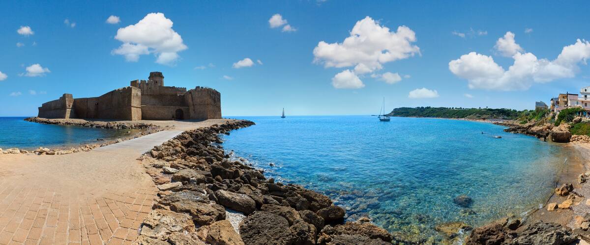 A coastal town with a historic stone fortress, clear blue waters, and a paved walkway along the shore.