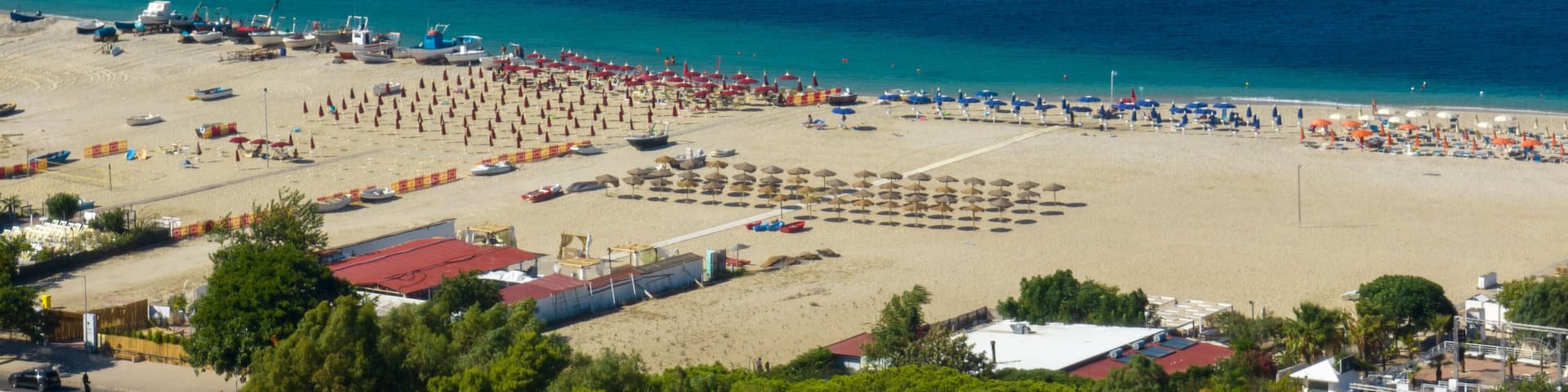 High-angle aerial view of a vibrant sandy beach featuring rows of organized beach umbrellas and sun loungers, bordered by lush green trees and the deep blue Mediterranean sea.