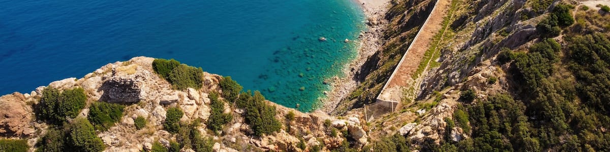 A coastal landscape with a winding road along the cliffside, a beach, and a clear view of the sea.