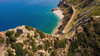 A coastal landscape with a winding road along the cliffside, a beach, and a clear view of the sea.