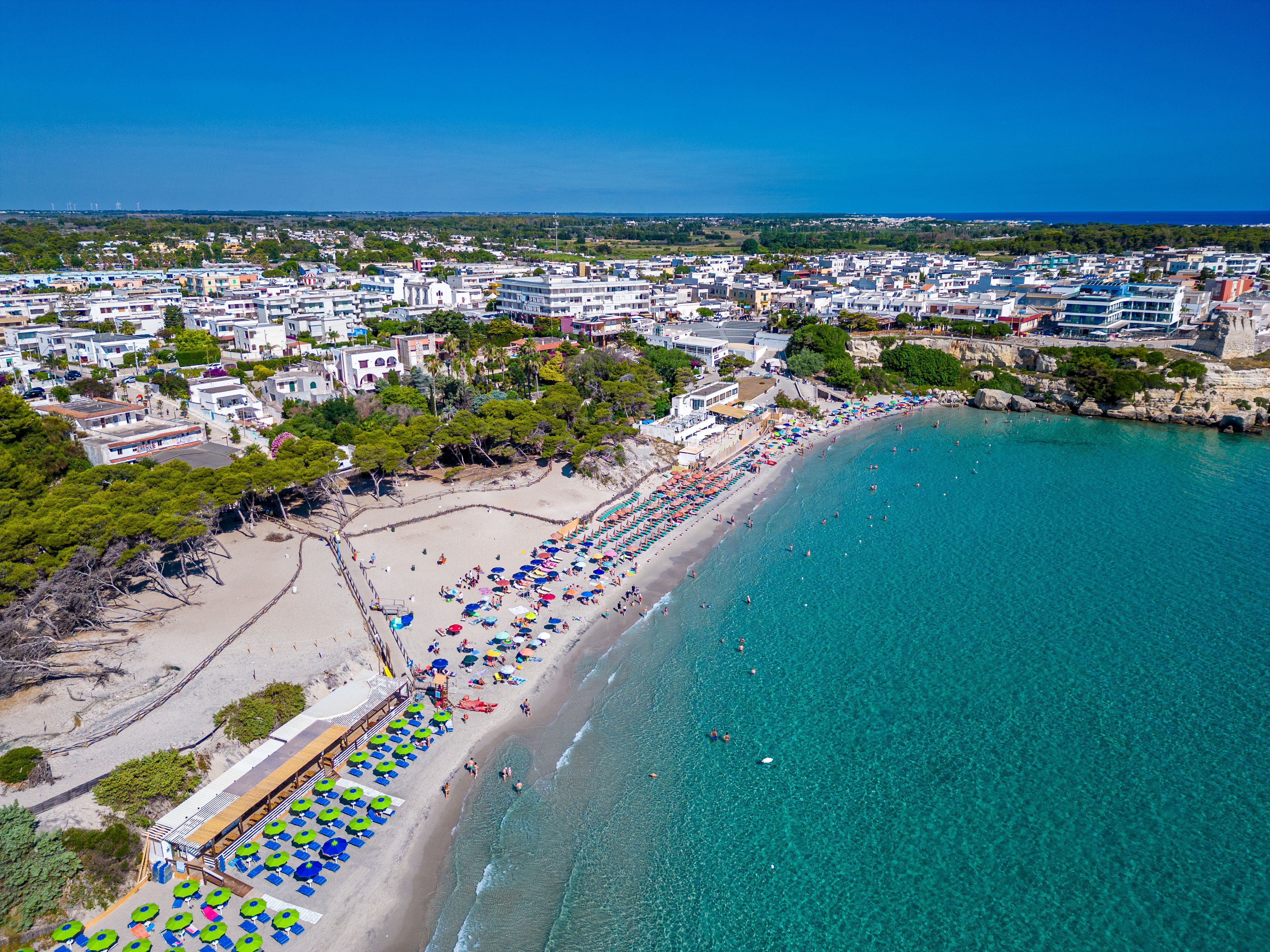 A coastal cityscape with a beach, clear turquoise waters, and a dense urban area with buildings and greenery.
