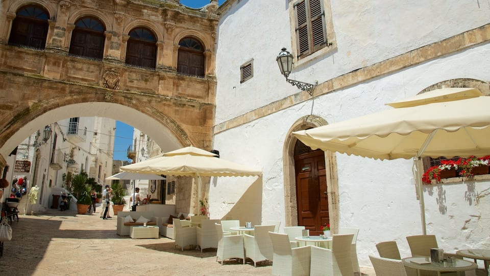 A historic street with white buildings, an archway, outdoor seating with umbrellas, and a café.