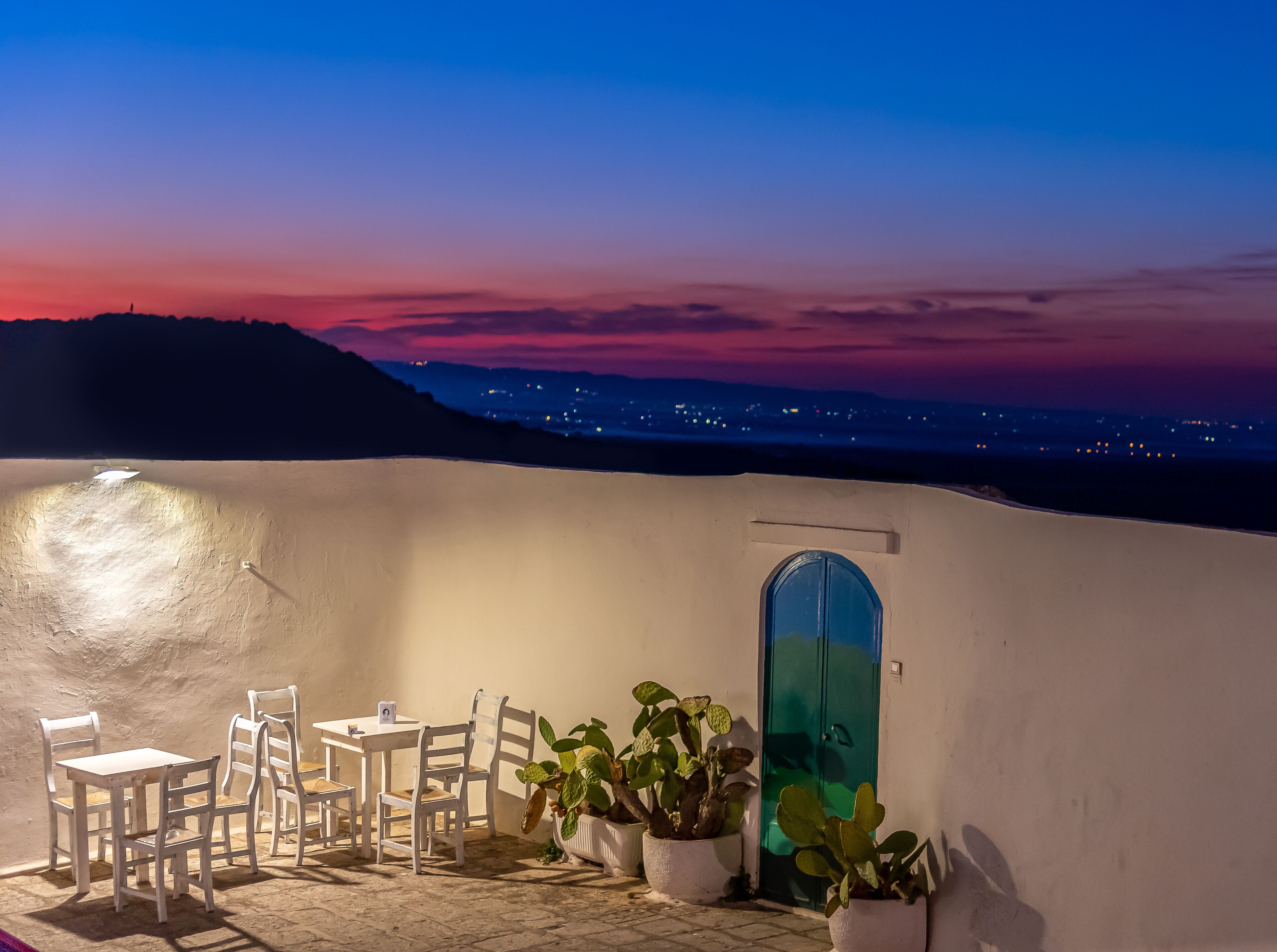 Una terraza de paredes blancas con una mesa, sillas, plantas en macetas y una puerta azul.