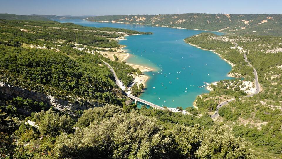 A coastal landscape with a turquoise bay, a bridge, and surrounding forested hills.