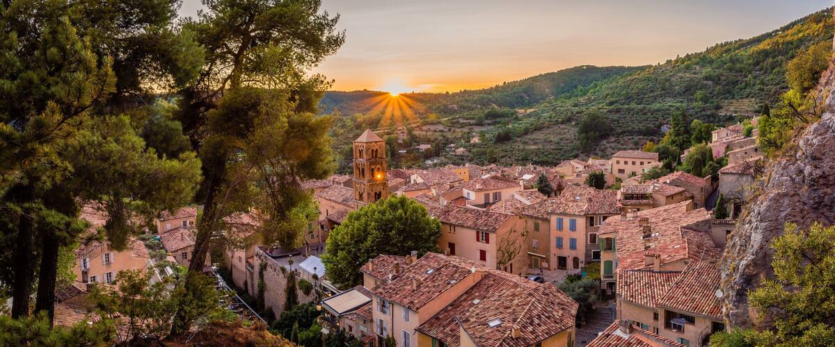 A village with terracotta-roofed buildings nestled in a valley, surrounded by trees and hills.