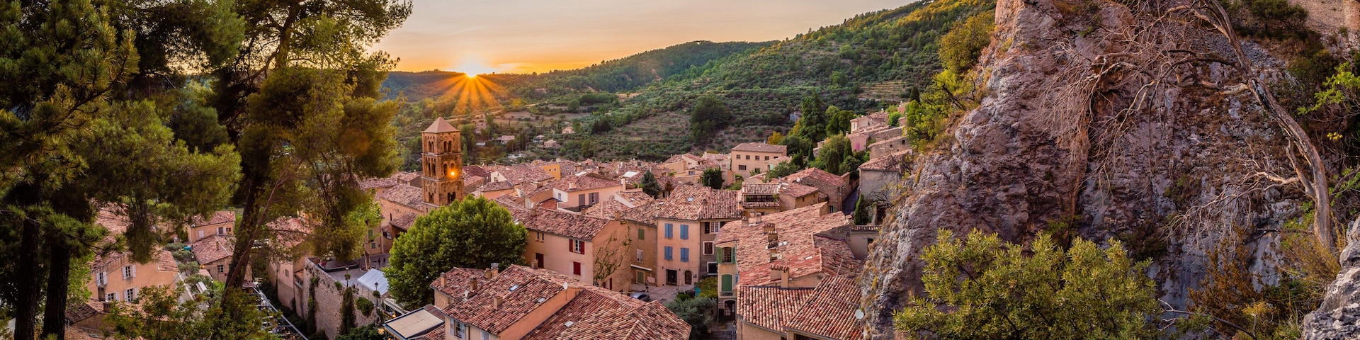 A village with terracotta-roofed buildings nestled in a valley, surrounded by trees and hills.