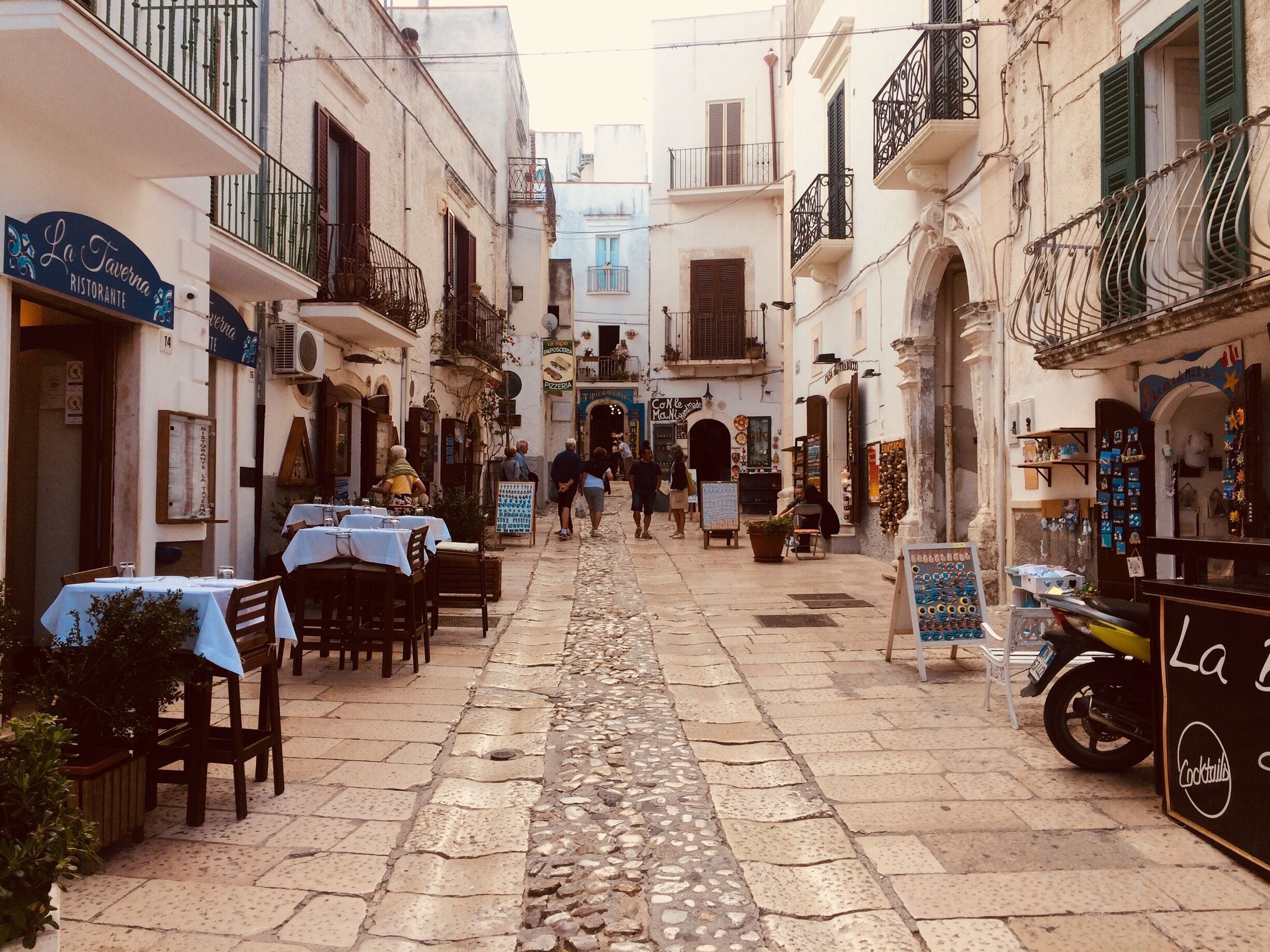 Une ruelle étroite pavée, bordée d’immeubles, avec des terrasses de café et une moto garée sur le bord.