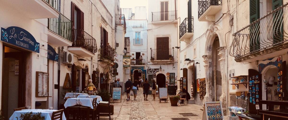 Una estrecha calle adoquinada bordeada de edificios, con mesas al aire libre en cafés y una motocicleta estacionada al costado.