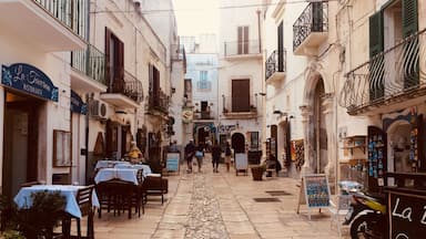 Uma rua de paralelepípedos estreita, ladeada por edifícios e com mesas de café ao ar livre, além de uma motocicleta estacionada na lateral.
