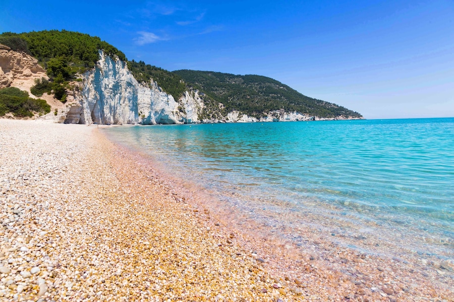 A pebble beach with clear turquoise water, white cliffs, and green hills.