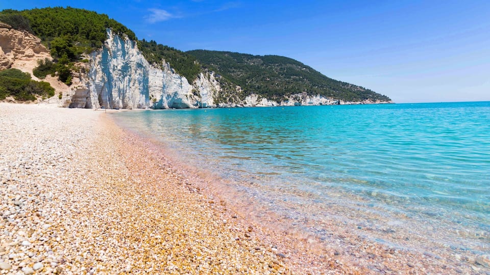 A pebble beach with clear turquoise water, white cliffs, and green hills.