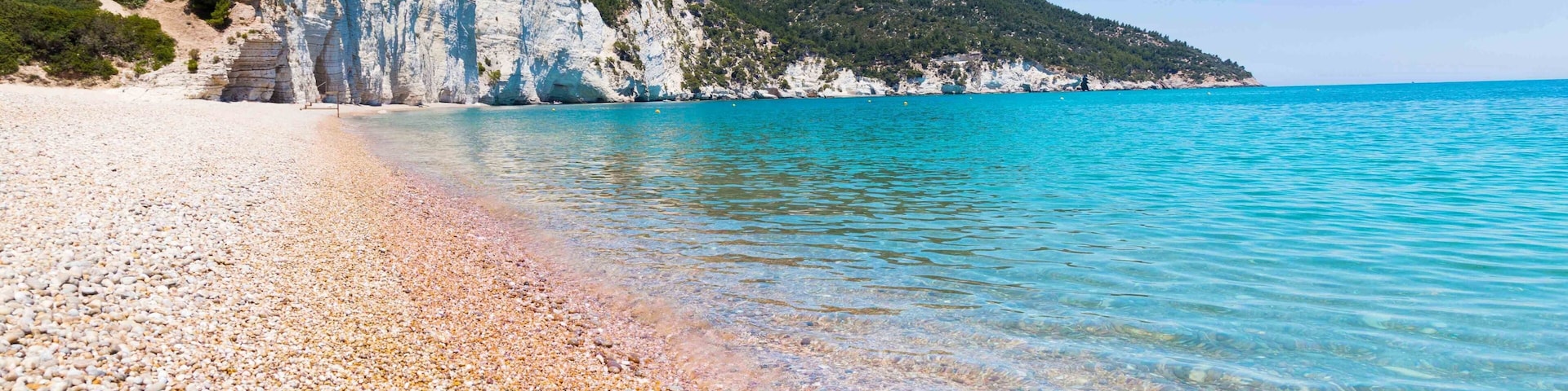 A pebble beach with clear turquoise water, white cliffs, and green hills.