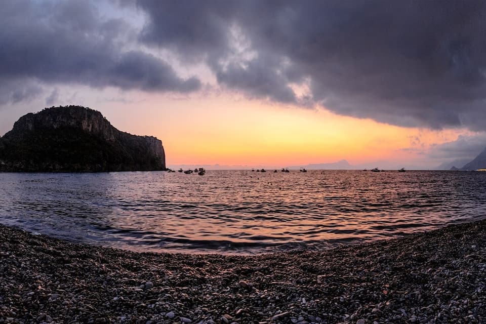 A pebbly beach with a boat, a rocky island, and a sunset over the water.