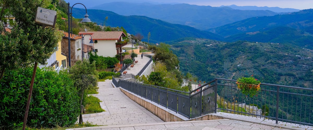 A paved pathway leading uphill with a view of a village and mountains in the distance.