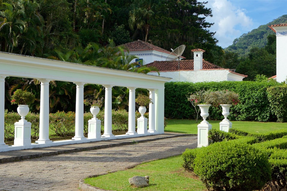 A white pavilion with columns, a garden with trimmed bushes, and a house with a red-tiled roof.