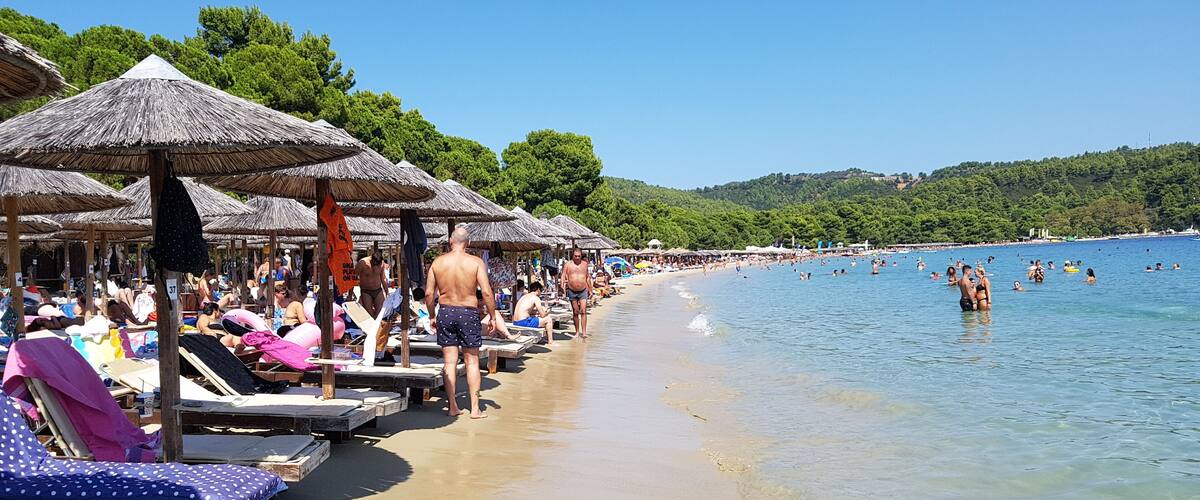 A beach with thatched umbrellas, sun loungers, and people swimming in the water.