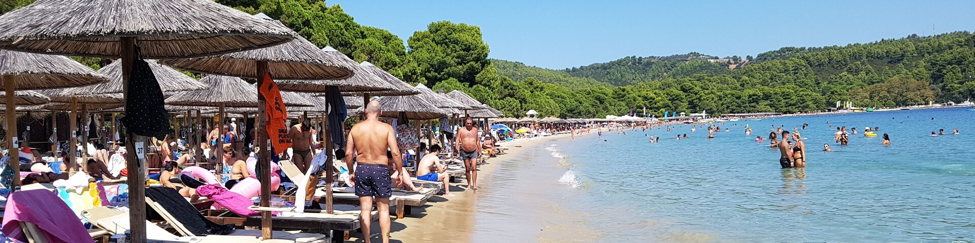 A beach with thatched umbrellas, sun loungers, and people swimming in the water.