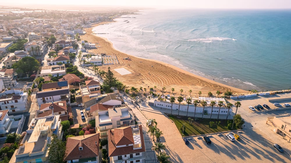 An aerial view of a coastal town with a beach, residential buildings, and parked cars.