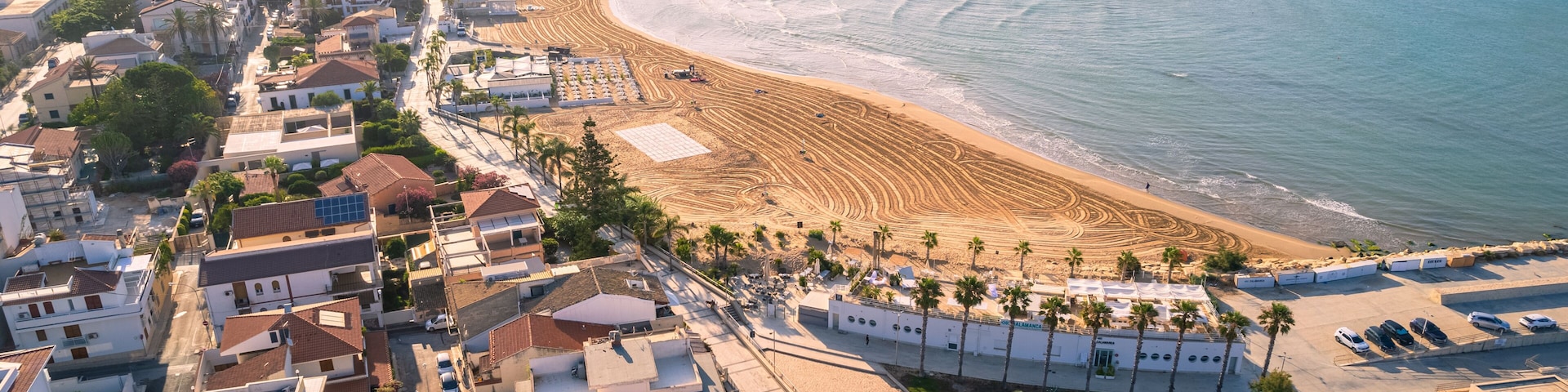An aerial view of a coastal town with a beach, residential buildings, and parked cars.