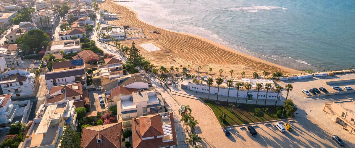 An aerial view of a coastal town with a beach, residential buildings, and parked cars.