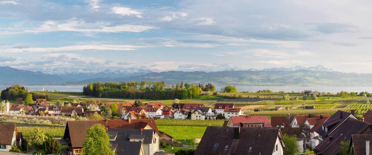 A village with red-roofed houses, vineyards, and a lake with mountains in the background.