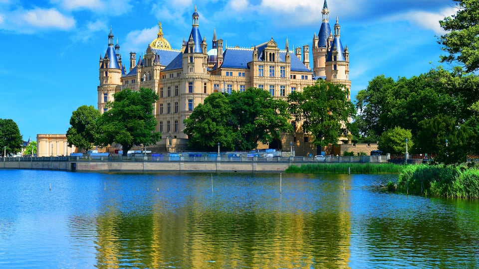 A grand castle with multiple towers and spires, a large body of water in the foreground, and lush green trees surrounding the castle.